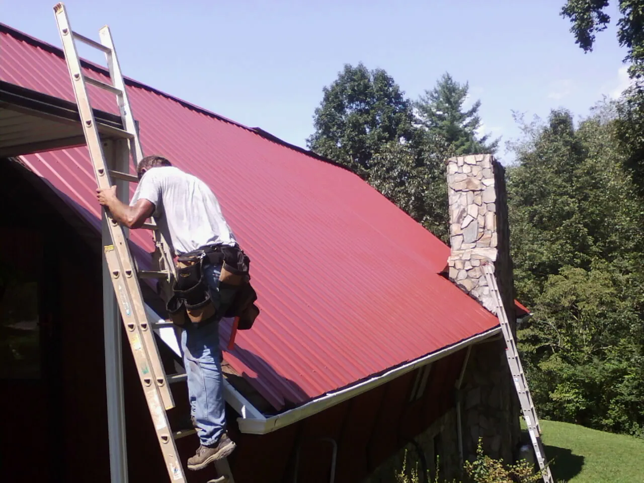 Roofer on a ladder beside a red metal roof and stone chimney — Soffit & Fascia Repair by Roofing Brothers in Maupin